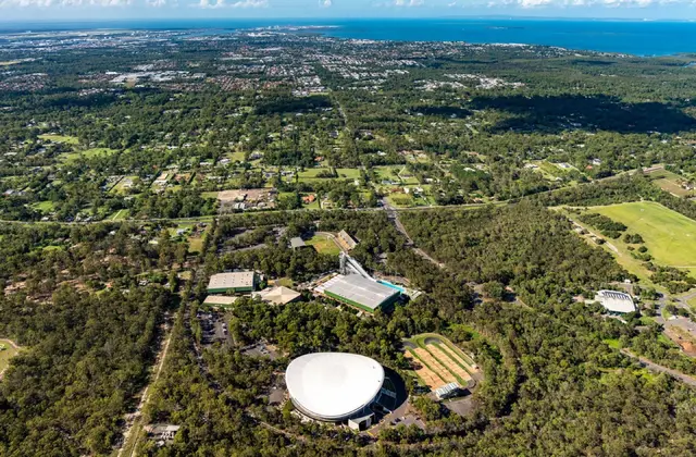 Aerial view of a large white domed velodrome ringed by car parks, with green-roofed aquatic and indoor sports buildings, a diving tower and a dirt BMX track alongside, all set in dense bushland; suburban houses, a golf course and the bay stretch to the horizon under scattered cloud.
