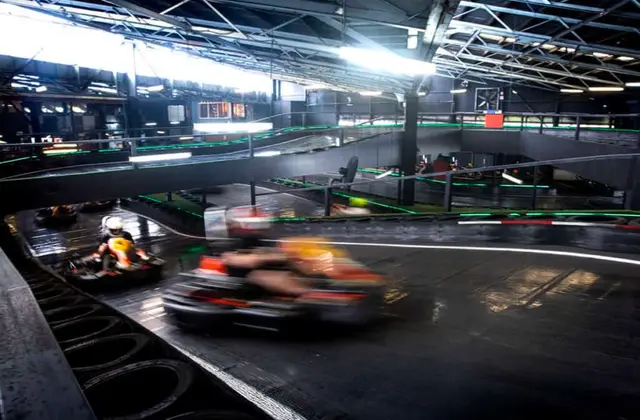 Wide view of the indoor kart track from above: two karts race along the dark lower level while a second storey of track, edged in green lights and steel railings, loops overhead beneath an exposed warehouse roof. Stacked tyre barriers line the inside of the bend.