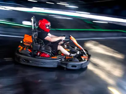 A driver in a red full-face helmet and black t-shirt leans into a corner in an orange and black go kart, the indoor track's green LED strip lighting blurred into streaks behind them.