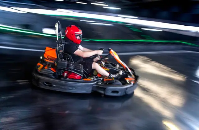 A driver in a red full-face helmet and black t-shirt leans into a corner in an orange and black go kart, the indoor track's green LED strip lighting blurred into streaks behind them.