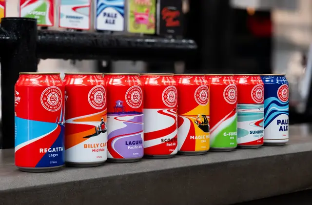 Eight 375ml beer cans lined up along a grey concrete bar top, each carrying a circular brewery roundel and swooping racing-stripe artwork in a different colour, labelled Regatta Lager, Billy Cart Mid Pale, Laguna Pacific Ale, Scooter Red Ale, Magic Mile XPA, G-Force IPA, Sundrift Hazy XPA and Pale Ale; tap badges hang on a black rail behind them.