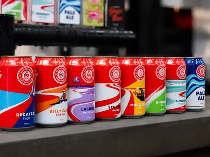 Eight 375ml beer cans lined up along a grey concrete bar top, each carrying a circular brewery roundel and swooping racing-stripe artwork in a different colour, labelled Regatta Lager, Billy Cart Mid Pale, Laguna Pacific Ale, Scooter Red Ale, Magic Mile XPA, G-Force IPA, Sundrift Hazy XPA and Pale Ale; tap badges hang on a black rail behind them.