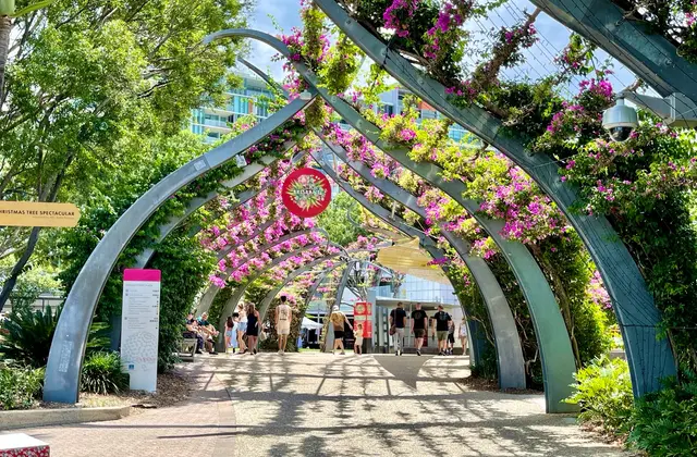 The Grand Arbour walkway — a long run of curved steel arches covered in flowering magenta bougainvillea — with people strolling the shaded path beneath it.