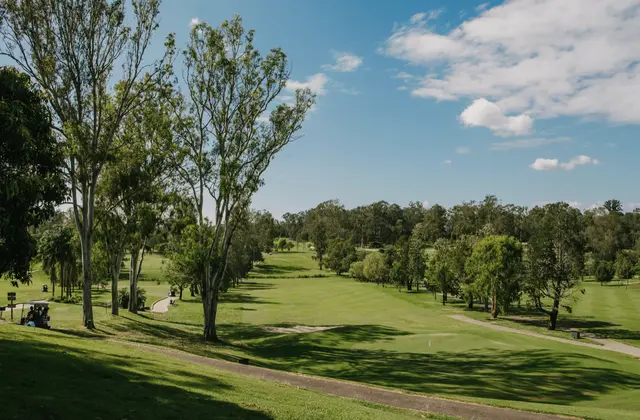 The course looking down a fairway from a grassy rise: tall pale-trunked eucalypts on the left throwing long shadows across mown grass, a concrete cart path curving through, a green with a flagstick at lower right, and a wall of bushland trees along the far side under a blue sky with scattered cloud. Two golf carts are parked in the shade at the left.