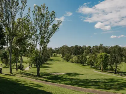 The course looking down a fairway from a grassy rise: tall pale-trunked eucalypts on the left throwing long shadows across mown grass, a concrete cart path curving through, a green with a flagstick at lower right, and a wall of bushland trees along the far side under a blue sky with scattered cloud. Two golf carts are parked in the shade at the left.