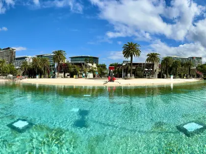 The man-made swimming lagoon photographed from water level: clear turquoise water over a pale sand shelf, a beach fringed with palms on the far side, a lifeguard in red standing on the sand and apartment towers behind.