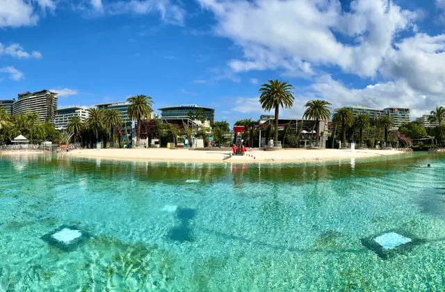 The man-made swimming lagoon photographed from water level: clear turquoise water over a pale sand shelf, a beach fringed with palms on the far side, a lifeguard in red standing on the sand and apartment towers behind.