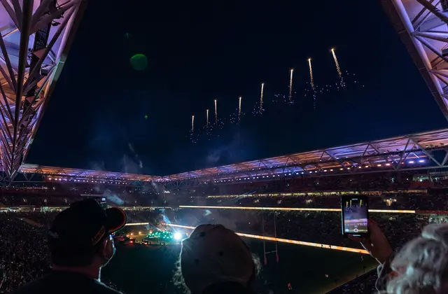 A night view from high in the stands: spectators in caps in the foreground, one holding up a phone to film, as fireworks trail up through the open gap in the roof above a floodlit green field, packed tiers of seating and lit signage ringing the ground.