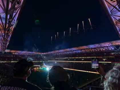 A night view from high in the stands: spectators in caps in the foreground, one holding up a phone to film, as fireworks trail up through the open gap in the roof above a floodlit green field, packed tiers of seating and lit signage ringing the ground.