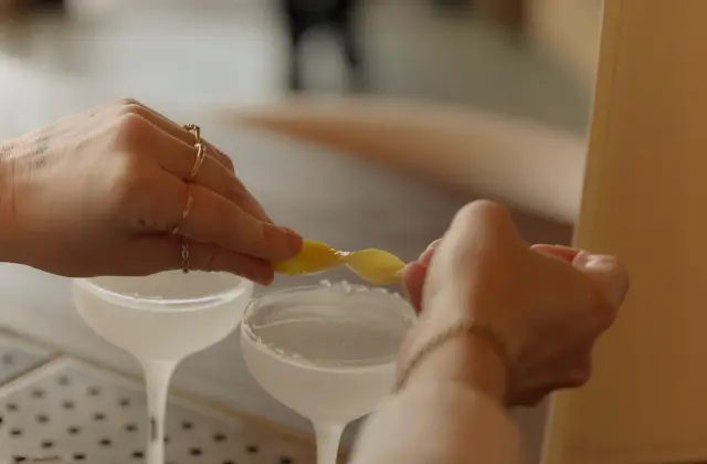 Two coupe glasses of a pale clear cocktail sit on a perforated metal bar mat while one person twists a strip of lemon peel over them and another steadies a glass; a folded card menu stands behind.