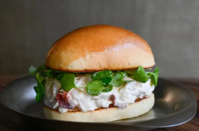 A glossy soft milk bun split and filled with a thick creamy seafood salad and a tangle of watercress, served on a small stainless steel plate on a dark timber table.