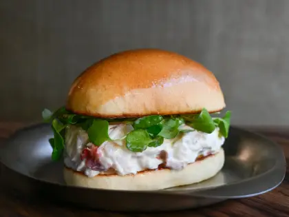 A glossy soft milk bun split and filled with a thick creamy seafood salad and a tangle of watercress, served on a small stainless steel plate on a dark timber table.