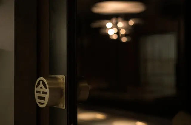 A dark timber-framed glass door standing ajar, fitted with a round brass handle plate engraved with a stylised character, opening onto a dimly lit dining room where a cluster of small warm pendant lights hangs above.