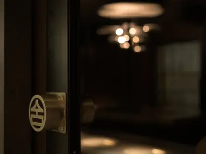 A dark timber-framed glass door standing ajar, fitted with a round brass handle plate engraved with a stylised character, opening onto a dimly lit dining room where a cluster of small warm pendant lights hangs above.