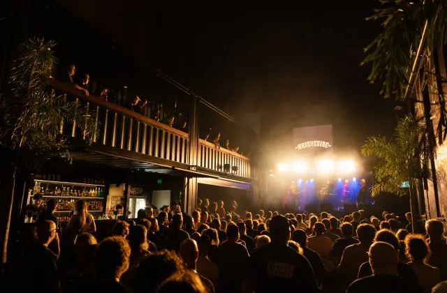 A dense standing crowd watches a four-piece band play under white and blue stage lights at night, with a lit screen above the stage showing The Brightside logo, a spirits back bar at the left and more people leaning over an upper-level balcony rail.
