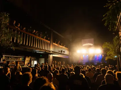 A dense standing crowd watches a four-piece band play under white and blue stage lights at night, with a lit screen above the stage showing The Brightside logo, a spirits back bar at the left and more people leaning over an upper-level balcony rail.