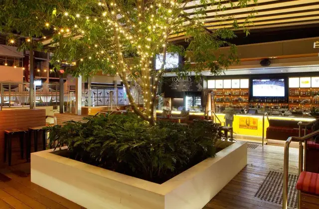 The beer garden at night: a tree strung with fairy lights growing from a raised white planter box on a timber deck, gas patio heaters and bar stools to one side, and a lit spirits bar with television screens and red banquette booths to the other.
