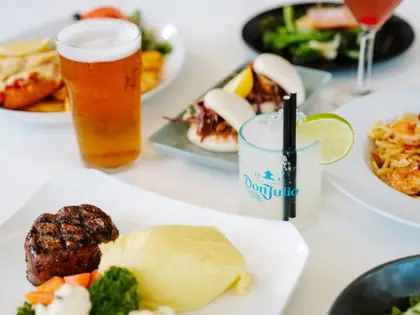A white table crowded with plates: a pint of amber beer, a tumbler of a clear iced drink garnished with a lime wheel, steamed bao buns with slaw and lemon, a fillet steak with mashed potato and vegetables, a prawn and chilli pasta, a plate of crumbed seafood, and a dish of greens with salmon; a pink cocktail in a stemmed glass sits at the back.