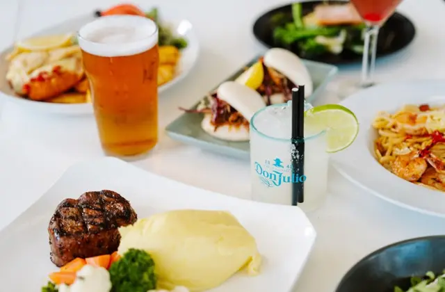 A white table crowded with plates: a pint of amber beer, a tumbler of a clear iced drink garnished with a lime wheel, steamed bao buns with slaw and lemon, a fillet steak with mashed potato and vegetables, a prawn and chilli pasta, a plate of crumbed seafood, and a dish of greens with salmon; a pink cocktail in a stemmed glass sits at the back.