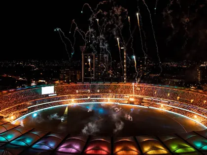 Aerial night view of a round stadium whose segmented roof canopy is lit in bands of pink, orange, green and blue; the tiers are packed with spectators, smoke and coloured light drift across the floodlit oval below, and fireworks trail overhead against a dark skyline of apartment towers and a construction crane.