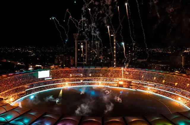Aerial night view of a round stadium whose segmented roof canopy is lit in bands of pink, orange, green and blue; the tiers are packed with spectators, smoke and coloured light drift across the floodlit oval below, and fireworks trail overhead against a dark skyline of apartment towers and a construction crane.