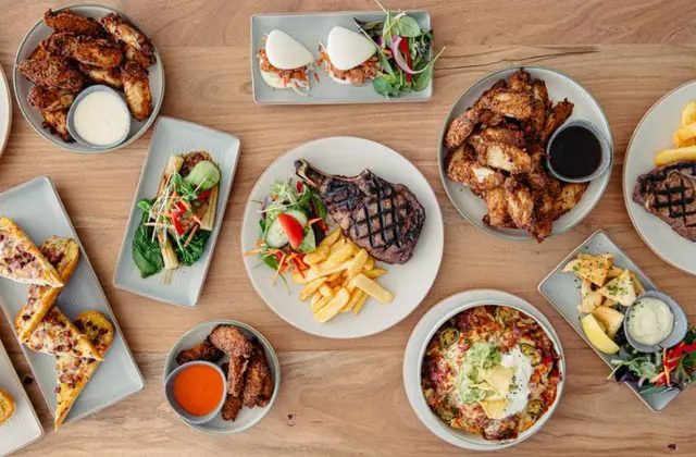 Overhead view of pub dishes spread across a pale timber table: two bowls of fried chicken wings with pale and dark dipping sauces, a grilled tomahawk steak with chips and garden salad, sliders with slaw and leaves, a bowl of nachos topped with sour cream, guacamole and corn chips, slices of cheese-and-bacon topped bread, loaded potato pieces with aioli, and a small pot of hot sauce.