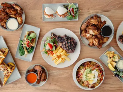 Overhead view of pub dishes spread across a pale timber table: two bowls of fried chicken wings with pale and dark dipping sauces, a grilled tomahawk steak with chips and garden salad, sliders with slaw and leaves, a bowl of nachos topped with sour cream, guacamole and corn chips, slices of cheese-and-bacon topped bread, loaded potato pieces with aioli, and a small pot of hot sauce.