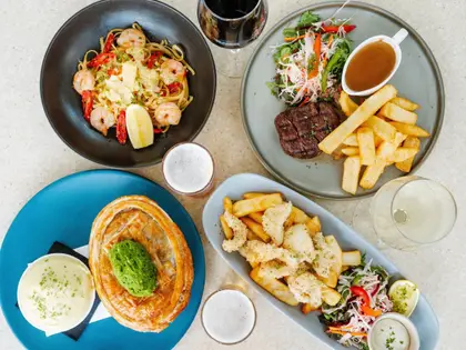 Overhead view of four meals on a speckled cream tabletop: prawn linguine with chilli and lemon in a black bowl, a steak with thick chips, salad and a jug of gravy, a pastry-topped pie with a spoonful of mushy peas beside a bowl of mash, and salt-and-pepper calamari with chips, salad and aioli; a glass of red wine, a glass of white and two glasses of beer stand between the plates.