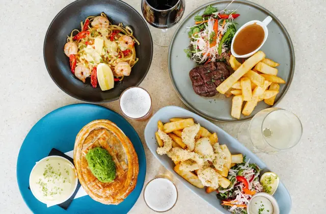 Overhead view of four meals on a speckled cream tabletop: prawn linguine with chilli and lemon in a black bowl, a steak with thick chips, salad and a jug of gravy, a pastry-topped pie with a spoonful of mushy peas beside a bowl of mash, and salt-and-pepper calamari with chips, salad and aioli; a glass of red wine, a glass of white and two glasses of beer stand between the plates.
