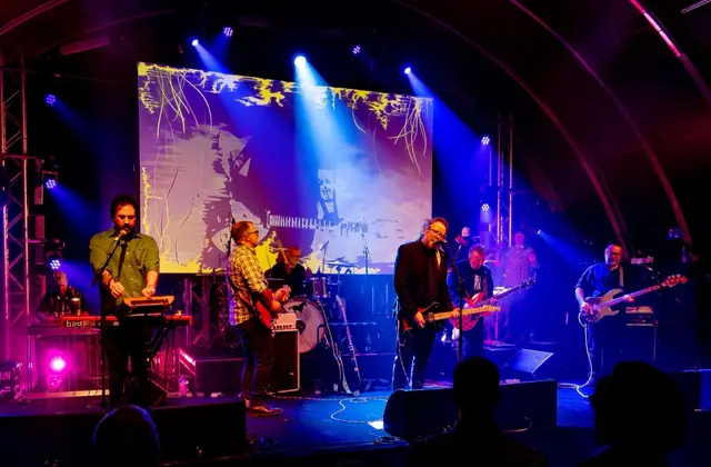 A band playing on stage under blue and magenta stage lights, a projected abstract backdrop behind them and the curved steel ribs of the hangar roof arching overhead above the heads of the audience.