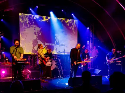 A band playing on stage under blue and magenta stage lights, a projected abstract backdrop behind them and the curved steel ribs of the hangar roof arching overhead above the heads of the audience.