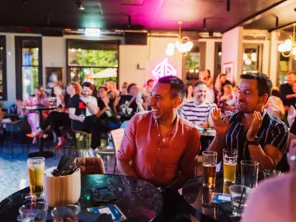 Two seated guests laugh and applaud at a marble-topped table holding glasses of beer, with a full room of patrons behind them and a pink-lit neon sign and pendant lights overhead.
