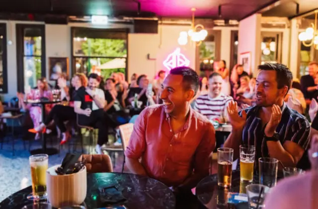 Two seated guests laugh and applaud at a marble-topped table holding glasses of beer, with a full room of patrons behind them and a pink-lit neon sign and pendant lights overhead.