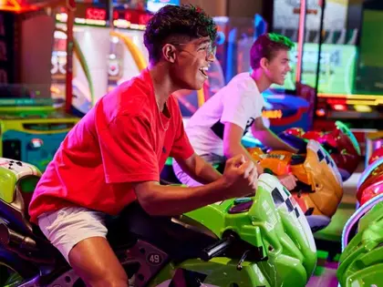 Two teenagers laughing as they lean over the handlebars of green and orange motorbike racing cabinets in an arcade, one punching the air, surrounded by brightly lit game machines, neon-edged screens and a patterned carpet.