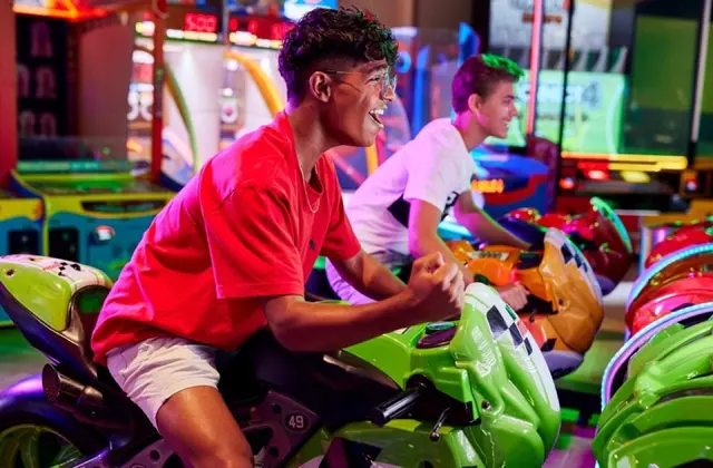 Two teenagers laughing as they lean over the handlebars of green and orange motorbike racing cabinets in an arcade, one punching the air, surrounded by brightly lit game machines, neon-edged screens and a patterned carpet.