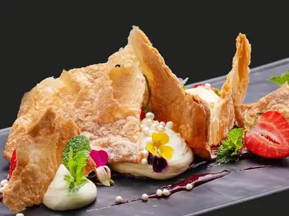 A plated dessert on a long slate-grey dish against a black background: shards of flaky puff pastry dusted with icing sugar propped between quenelles of cream, a smear of dark berry sauce, small white sugar pearls, halved strawberries, mint leaves and purple and yellow edible violas.