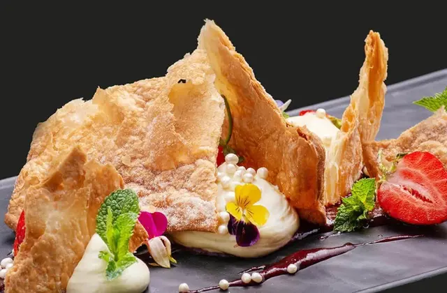 A plated dessert on a long slate-grey dish against a black background: shards of flaky puff pastry dusted with icing sugar propped between quenelles of cream, a smear of dark berry sauce, small white sugar pearls, halved strawberries, mint leaves and purple and yellow edible violas.