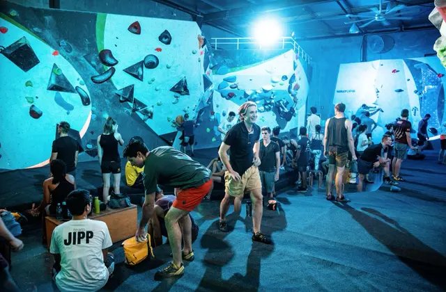 A busy bouldering hall lit blue-green: tall pale angled walls studded with brightly coloured holds run down both sides under a dark corrugated roof with ceiling fans, and about twenty climbers in shorts and t-shirts stand, sit on benches or crouch to change shoes on the black matted floor while one climber in the centre stretches out mid-conversation.