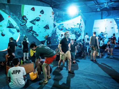 A busy bouldering hall lit blue-green: tall pale angled walls studded with brightly coloured holds run down both sides under a dark corrugated roof with ceiling fans, and about twenty climbers in shorts and t-shirts stand, sit on benches or crouch to change shoes on the black matted floor while one climber in the centre stretches out mid-conversation.