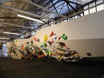 A long overhanging bouldering wall inside a warehouse: a smooth cream-painted prow angles out over black matting, its underside covered in green, red, yellow, blue and white holds, with fluorescent strip lights and an exposed steel truss roof above and a small figure standing at the far end of the empty floor.