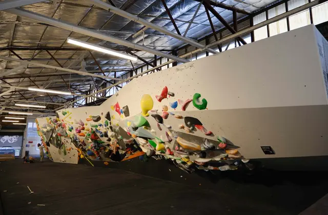 A long overhanging bouldering wall inside a warehouse: a smooth cream-painted prow angles out over black matting, its underside covered in green, red, yellow, blue and white holds, with fluorescent strip lights and an exposed steel truss roof above and a small figure standing at the far end of the empty floor.