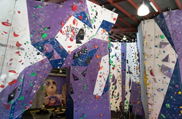 The climbing hall seen from the floor: tall purple, navy and white angled walls studded with red, green, orange and blue holds rise toward a red steel truss roof. Ropes hang down the taller walls on the right where two climbers are tying in, and a large mural of a child in orange-rimmed sunglasses is painted on the far wall.