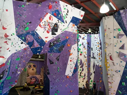 The climbing hall seen from the floor: tall purple, navy and white angled walls studded with red, green, orange and blue holds rise toward a red steel truss roof. Ropes hang down the taller walls on the right where two climbers are tying in, and a large mural of a child in orange-rimmed sunglasses is painted on the far wall.