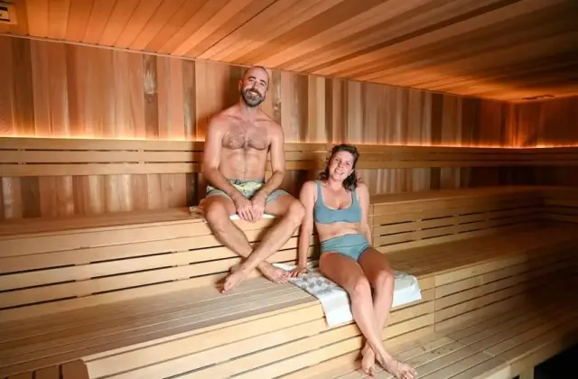 Inside the sauna: tiered pale timber benches under a slatted timber ceiling, warm strip lighting running along the wall behind the top bench. A man in board shorts sits on the middle tier with his feet on the bench below; a woman in a grey-blue two-piece reclines beside him on a folded towel. Both are smiling.