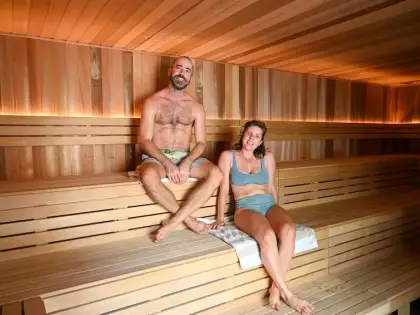 Inside the sauna: tiered pale timber benches under a slatted timber ceiling, warm strip lighting running along the wall behind the top bench. A man in board shorts sits on the middle tier with his feet on the bench below; a woman in a grey-blue two-piece reclines beside him on a folded towel. Both are smiling.