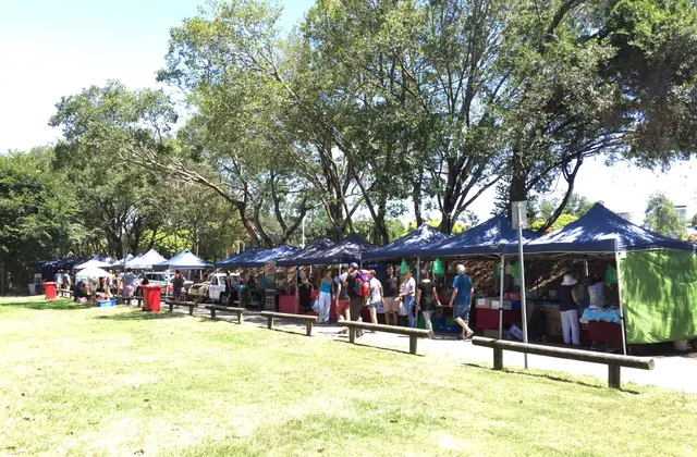 A row of navy market marquees pitched on grass under big shade trees, with shoppers walking the line of stalls past produce crates and trestle tables.