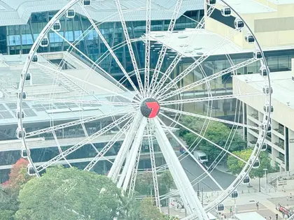 The white observation wheel seen from above, its ring of enclosed dark gondolas turning around a hub marked with a red Channel Seven logo, with the tented white roofs of the convention centre behind and treetops below.