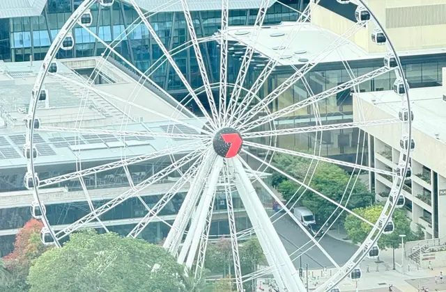 The white observation wheel seen from above, its ring of enclosed dark gondolas turning around a hub marked with a red Channel Seven logo, with the tented white roofs of the convention centre behind and treetops below.