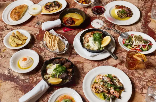 Overhead view of a shared meal spread across a red-veined marble table: charred octopus, a skillet of fried cheese with chilli jam, lemon-roasted potatoes, warm flatbreads in a wire basket, a dish of dip topped with orange roe, a skillet of chicken and rice with dill and lemon, a plate of braised meat over rice, grilled prawns in oil with spring onion, and a salad of tomato, cucumber and onion under a slab of feta; a glass of red wine, a spirit cocktail with dried orange and a small round table marker reading Yamas sit among them.