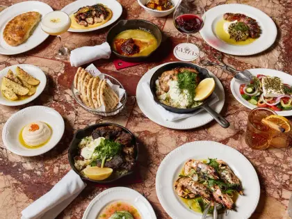 Overhead view of a shared meal spread across a red-veined marble table: charred octopus, a skillet of fried cheese with chilli jam, lemon-roasted potatoes, warm flatbreads in a wire basket, a dish of dip topped with orange roe, a skillet of chicken and rice with dill and lemon, a plate of braised meat over rice, grilled prawns in oil with spring onion, and a salad of tomato, cucumber and onion under a slab of feta; a glass of red wine, a spirit cocktail with dried orange and a small round table marker reading Yamas sit among them.